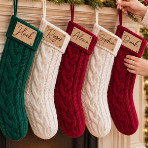 Puede incluir: Cinco calcetines navideños de punto trenzado en verde, blanco y rojo. Cada calcetín presenta una etiqueta rectangular de aspecto cuero sujeta con remaches metálicos, personalizada con los nombres Hunk, Roger, Alexia, Sophia y Dinah en cursiva.