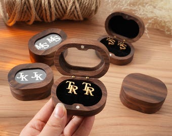 May include: Wooden jewellery boxes with personalised initials. The boxes are oval-shaped with a dark wood finish and a clear top. Inside, the boxes have a black velvet lining and hold gold-coloured initial earrings. The initials are visible in the boxes.