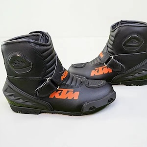 Könnte beinhalten: Paar schwarze Motorradstiefel mit orangefarbenem "KTM"-Branding. Die Stiefel haben ein hohes Design mit verstellbaren Riemen und verstärkten Einsätzen. Die Sohlen sind schwarz und strukturiert.