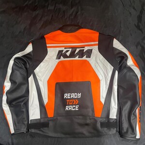 May include: A black, white, and orange motorcycle jacket with the KTM logo on the back. The jacket has a leather collar and the words "READY TO RACE" printed on the lower back.
