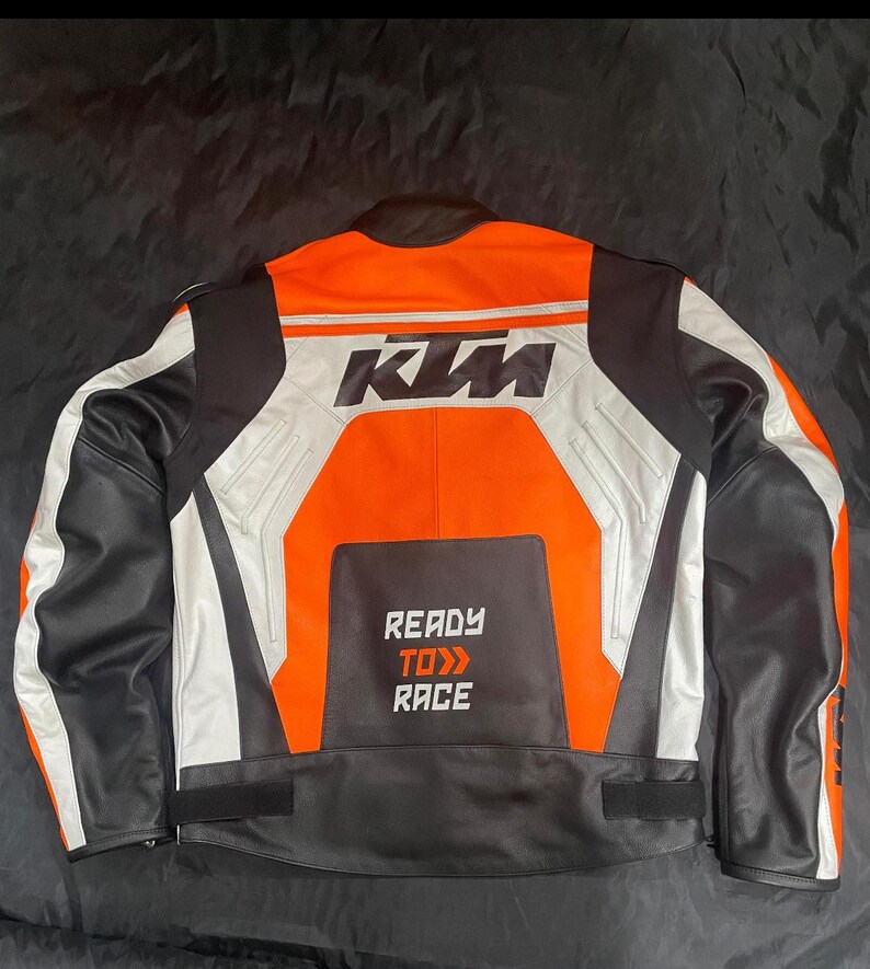 May include: A black, white, and orange motorcycle jacket with the KTM logo on the back. The jacket has a leather collar and the words "READY TO RACE" printed on the lower back.