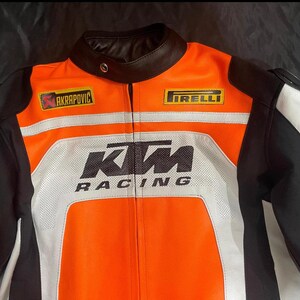 May include: Orange, black, and white motorcycle racing jacket with the KTM Racing logo. The jacket features the Akrapovic and Pirelli logos, a black leather collar, and white stripes. The jacket is designed for protection and speed.