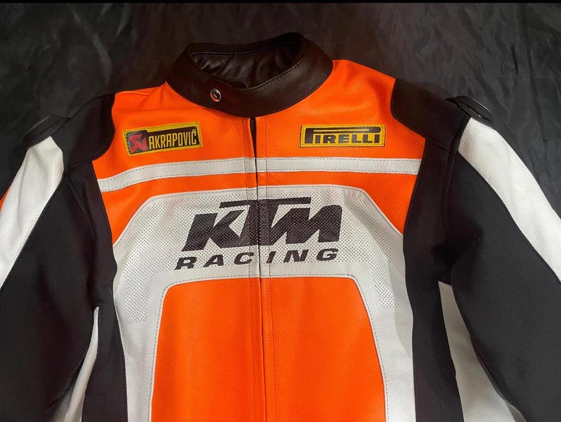 May include: Orange, black, and white motorcycle racing jacket with the KTM Racing logo. The jacket features the Akrapovic and Pirelli logos, a black leather collar, and white stripes. The jacket is designed for protection and speed.