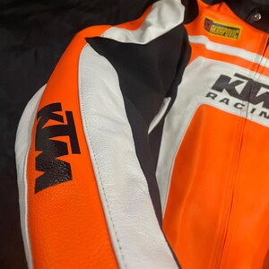 May include: Close-up of a motorcycle racing jacket. The jacket is primarily orange, with white and black accents. The KTM logo is prominently displayed on the sleeve. The jacket also features the Akrapovic logo and the words "KW RACING".