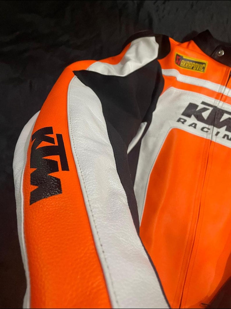 May include: Close-up of a motorcycle racing jacket. The jacket is primarily orange, with white and black accents. The KTM logo is prominently displayed on the sleeve. The jacket also features the Akrapovic logo and the words "KW RACING".