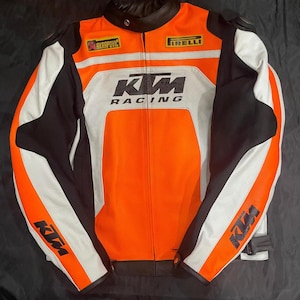 May include: Orange, black, and white racing jacket with the KTM Racing logo. The jacket features the Akrapovic and Pirelli logos, with white panels on the sleeves and a black collar.