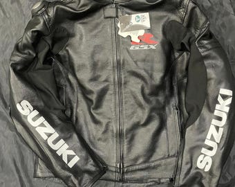 May include: Black leather motorcycle jacket with the word "SUZUKI" in white block letters on the sleeves. The jacket has a zipper closure and a small tag with the letters "GSX" in red. The jacket is made of leather and has a quilted lining.