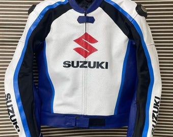 May include: White, blue, and black Suzuki motorcycle jacket. The jacket features the Suzuki logo in red and black on the chest, with blue and black accents on the sleeves and shoulders. The word "SUZUKI" is printed on the sleeves.