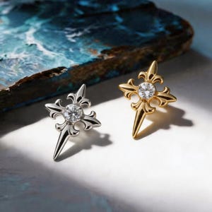 May include: Two stud earrings, one silver and one gold, each featuring a cross design with a central clear gemstone. The earrings have a fleur-de-lis style top and a pointed bottom. The background is a mix of blue and white.