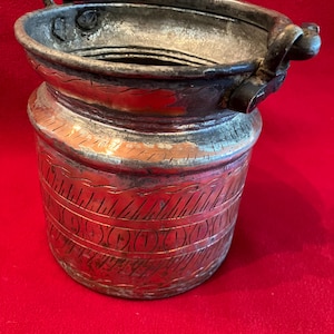 May include: A cylindrical metal bucket with a weathered silver and copper-toned finish. The exterior features horizontal bands of etched linear and oval patterns. It includes a sturdy, hinged metal handle attached to the top rim with rivets.