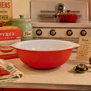 May include: A red and white Pyrex mixing bowl sits on a countertop next to a vintage Pyrex box, a flour canister, and a recipe card. A red spatula and measuring spoons are also visible, suggesting a baking theme.