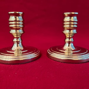 May include: A pair of polished brass candlesticks featuring a wide, circular tiered base and a central column with horizontal ribbed detailing. The candlesticks have a smooth, reflective metallic finish and a flared top rim for holding candles.