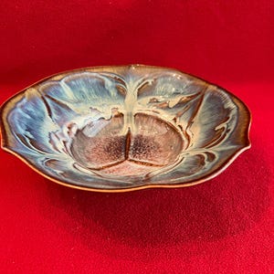 May include: A shallow ceramic bowl with a scalloped edge and a glossy glaze. The interior features a blend of deep brown, cream, and vibrant blue tones that radiate from the center toward the rim, creating a fluid, marbled, and multi-colored effect.