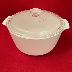 May include: A white round casserole dish with two integrated side handles and a clear glass lid featuring a central circular knob handle. The pot has a smooth, matte finish and a wide, tapered body design, providing a clean and functional appearance.