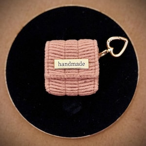 May include: A small, pink, handmade pouch with a rectangular gold-colored tag that reads "handmade." The pouch is attached to a gold-colored heart-shaped keychain. The pouch is sitting on a black circular surface.