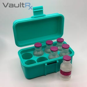 May include: A teal medical storage case with an open lid, holding six clear vials with pink caps. A single vial with a pink and white label sits outside the case. The VaultRx logo is visible in the upper left corner.