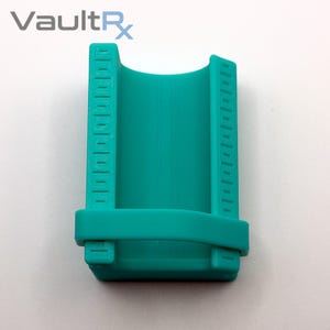 May include: A teal-colored, 3D-printed device with a curved interior and vertical measurement markings on the sides. The base has a rectangular shape with two small, square protrusions. The text "VaultRx" is in the upper left corner.