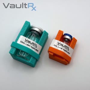 May include: Two label applicators, one teal and one orange, each holding a small glass vial. The teal applicator holds a 10mL vial, and the orange one holds a 3mL vial. Both applicators have the "VaultRx" logo and "Label Applicator" text.
