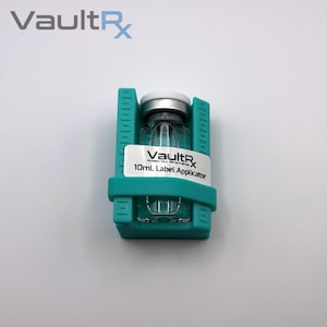 May include: A clear glass vial with a silver cap, held in a teal label applicator. The vial has a white label with the text "VaultRx 10mL Label Applicator". The VaultRx logo is visible at the top left.