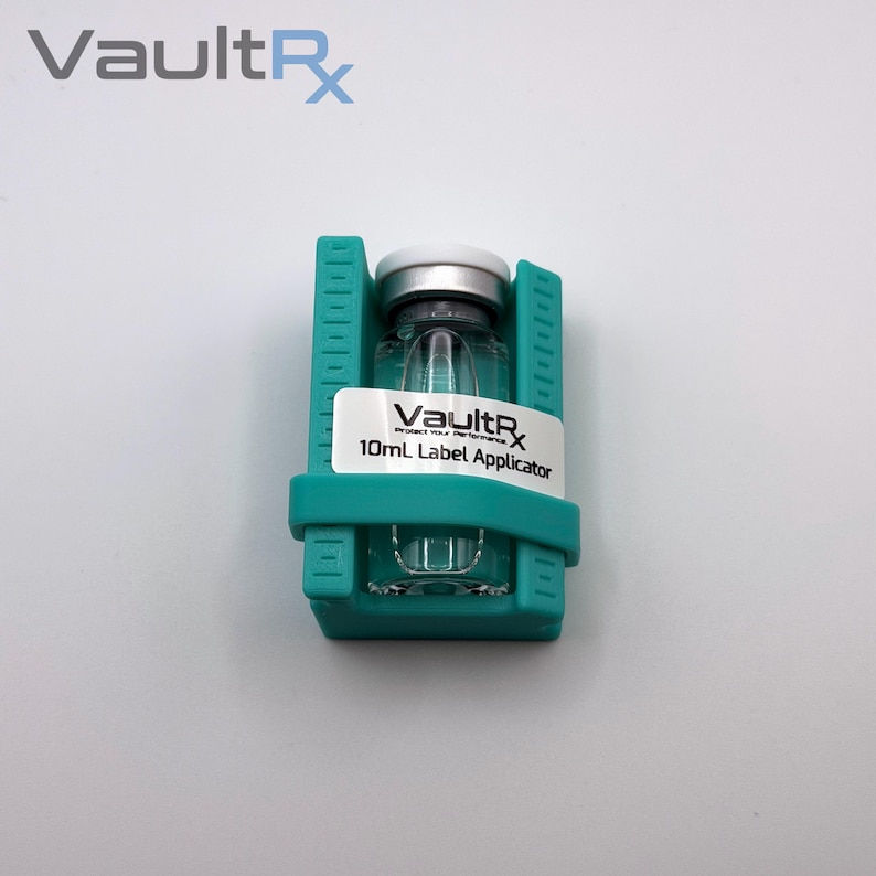 May include: A clear glass vial with a silver cap, held in a teal label applicator. The vial has a white label with the text "VaultRx 10mL Label Applicator". The VaultRx logo is visible at the top left.