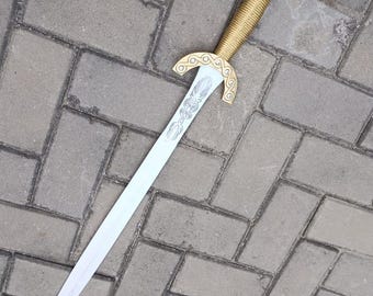 Puede incluir: Una espada decorativa con empuñadura y guarda doradas y una hoja plateada. La guarda presenta un diseño ornamentado. La espada está sobre una superficie de ladrillos grises.