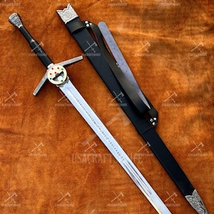 May include: A medieval sword with a silver blade and a black handle. The sword has a decorative crossguard and a black scabbard with silver detailing. The sword is displayed on a brown surface.