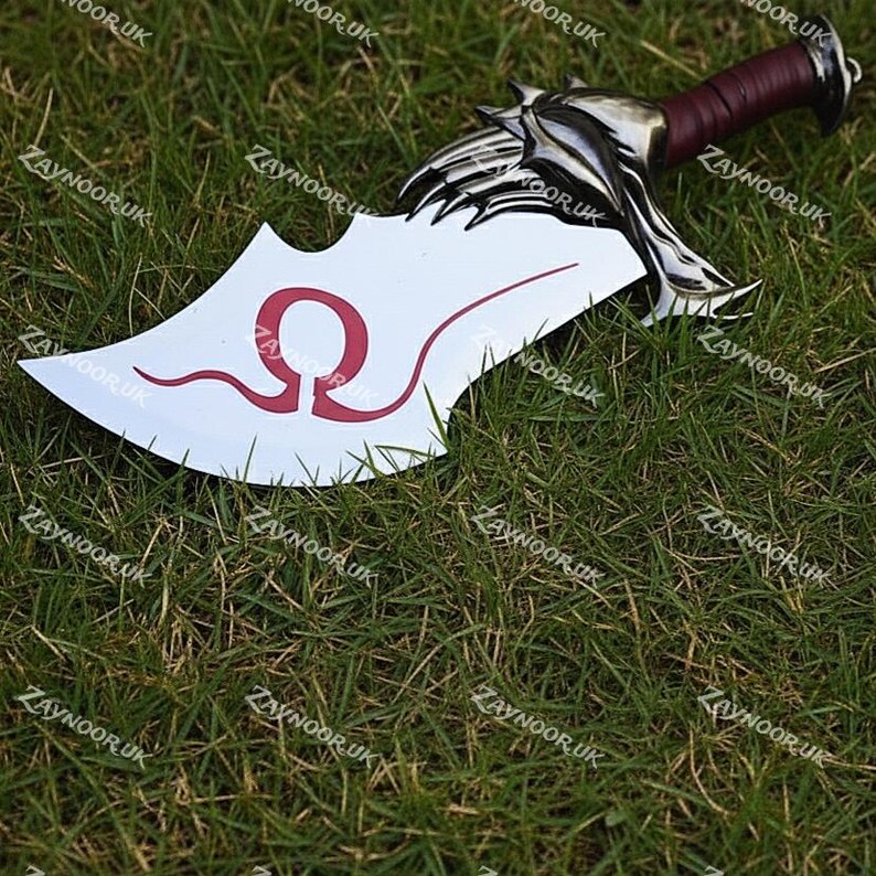 May include: A prop sword with a white blade featuring a red Omega symbol and a decorative silver hilt. The handle is wrapped in a dark red material. The sword is lying on a bed of green grass.