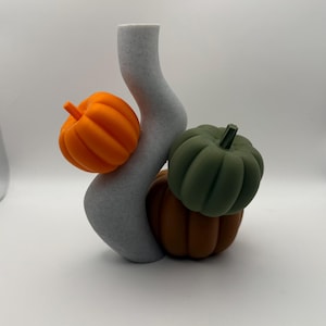 May include: A grey, wavy vase with an orange pumpkin attached, and two other pumpkins in shades of green and brown. The pumpkins have ribbed detailing and stems. The vase has a speckled texture and a smooth, curved shape.
