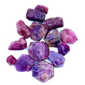 Pode incluir: Uma coleção de pedras preciosas de rubi brutas, não cortadas, em várias tonalidades de rosa e roxo. As pedras preciosas têm uma textura natural áspera e estão espalhadas sobre um fundo branco. As pedras variam em tamanho e forma.