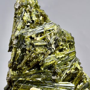 Op de afbeelding: Een cluster van doorschijnende, olijfgroene epidootkristallen met langwerpige, prismatische structuren en een glasachtige glans. Het mineraalexemplaar vertoont een complexe, in elkaar grijpende rangschikking van scherpe, naaldachtige formaties met kleine vlekken van lichtgekleurde matrix.
