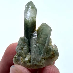 Puede incluir: Un grupo de cristales de cuarzo verde con una punta clara y afilada. Los cristales tienen una textura natural y rugosa y diferentes tonos de verde. El cuarzo se sostiene en una mano.