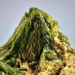 Op de afbeelding: Een ruw mineraal exemplaar met een cluster van levendige olijfgroene, naaldachtige kristallen die vanuit een centraal punt uitstralen. De kristallijne structuur is dicht en vezelig, geplaatst op een ruwe, poreuze, lichtbruine rotsachtige basis.