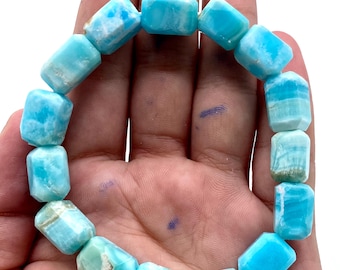 Può includere: Un braccialetto composto da perline rettangolari azzurre. Le perline hanno un aspetto marmorizzato con sfumature di bianco e verde. Il braccialetto è tenuto in una mano su sfondo bianco. Le perline misurano circa 2 cm di lunghezza.