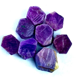 May include: A collection of raw, uncut ruby gemstones. The gemstones are a deep, rich purple color with visible crystalline structures and natural textures. The rubies are displayed against a white background.