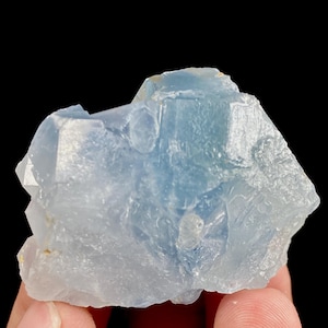 May include: A light blue, crystalline mineral specimen with a rough, textured surface. The mineral has a translucent quality, with variations in color from pale to medium blue. The specimen is held in a hand against a black background.