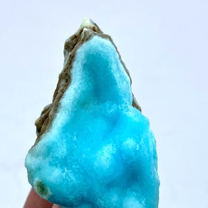 May include: A vibrant, natural mineral specimen featuring a striking combination of colors. The focal point is a vivid, light blue crystalline formation, contrasted by a rough, brown matrix. The mineral's surface has a textured appearance, with a mix of smooth and uneven areas.