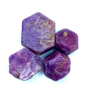 May include: Four rough-cut, natural ruby gemstones in varying sizes. The rubies display a deep, rich purple hue with visible surface textures and inclusions. The gemstones are set against a bright white background.