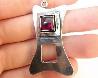May include: A silver pendant necklace with a geometric design. The pendant features a square, deep red gemstone set in a silver frame. The pendant has a rectangular cutout and is suspended from a delicate silver chain.