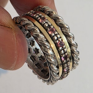May include: A stackable ring set featuring a silver-toned, twisted rope design. The set includes a gold-toned band with pink gemstones and a silver band with a cut-out pattern. The rings are held by a finger.