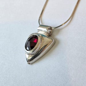 Puede incluir: Un collar con colgante de plata con un colgante triangular. El colgante presenta una gran piedra preciosa roja oscura de talla ovalada engastada en un bisel de plata. La cadena del collar es una delicada cadena de serpiente plateada.
