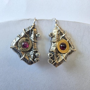 May include: A pair of artisan earrings. Each earring features a textured silver-tone metal design with gold-tone accents. A central, round, red gemstone is set within a gold circle. Small, clear gemstones are also set into the design.
