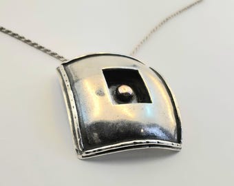 Puede incluir: Un collar con colgante plateado. El colgante tiene un recorte cuadrado con un pequeño acento redondo. El colgante tiene forma rectangular curva con un borde texturizado, suspendido de una delicada cadena.