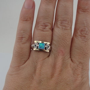 Op de afbeelding: Een zilveren ring met een vierkante, blauwgroene opaalsteen. De ring heeft gouden accenten en decoratieve krullen aan de zijkanten. De ring zit aan een hand.