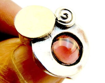 May include: A ring featuring a polished gold-tone circular disc, a silver-tone spiral accent, and a round, deep red cabochon stone set within a silver-tone metal base. The design combines mixed metal finishes with a prominent dark red gemstone.