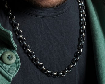Può includere: Primo piano di una persona che indossa una giacca verde scuro sopra una maglietta nera con una collana a catena argentata.