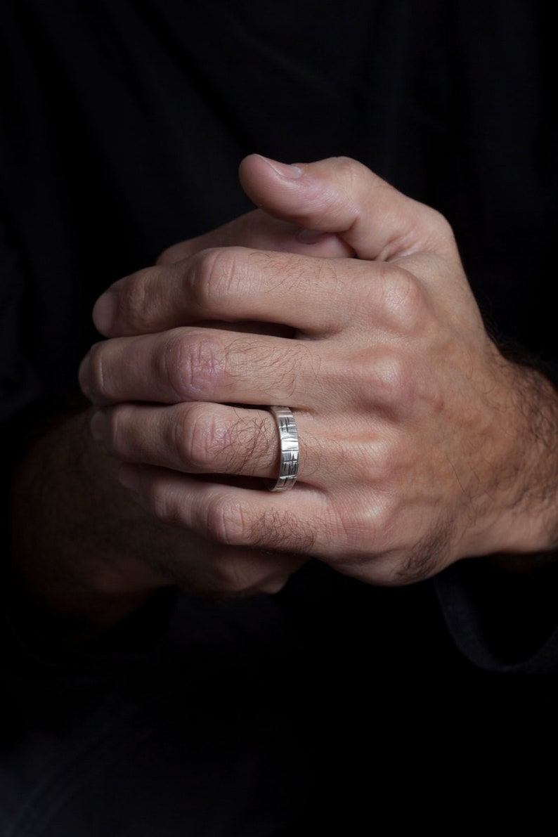 K&ouml;nnte beinhalten: Ein silberner Bandring mit einem strukturierten Design. Der Ring wird am Ringfinger der linken Hand einer Person getragen.