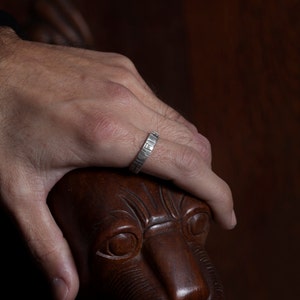K&ouml;nnte beinhalten: Ein silberner Ring mit einer strukturierten Band. Der Ring wird am Finger einer Hand getragen, die auf einem geschnitzten L&ouml;wenkopf aus Holz ruht.