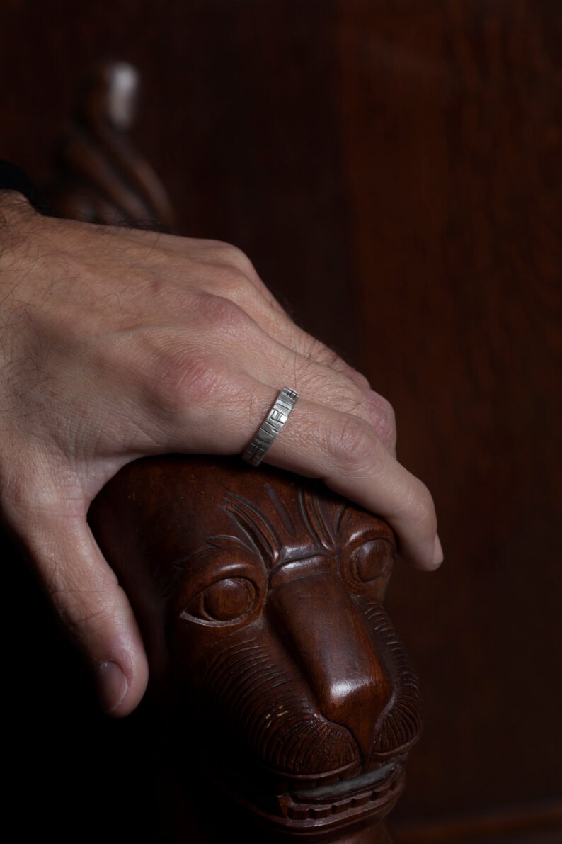 K&ouml;nnte beinhalten: Ein silberner Ring mit einer strukturierten Band. Der Ring wird am Finger einer Hand getragen, die auf einem geschnitzten L&ouml;wenkopf aus Holz ruht.