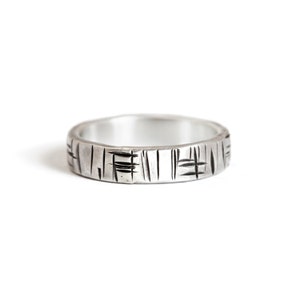 K&ouml;nnte beinhalten: Ein silberner Bandring mit einem strukturierten Design. Die Band ist glatt und gl&auml;nzend, mit einem Muster aus Linien und Strichen, die in die Oberfl&auml;che ge&auml;tzt sind.