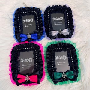 May include: Four ornate black picture frames with colorful accents. Each frame has a black velvet border, a ruffled trim in blue, purple, pink, and green, and a satin bow. The frames feature the text "hatiskoll designs".